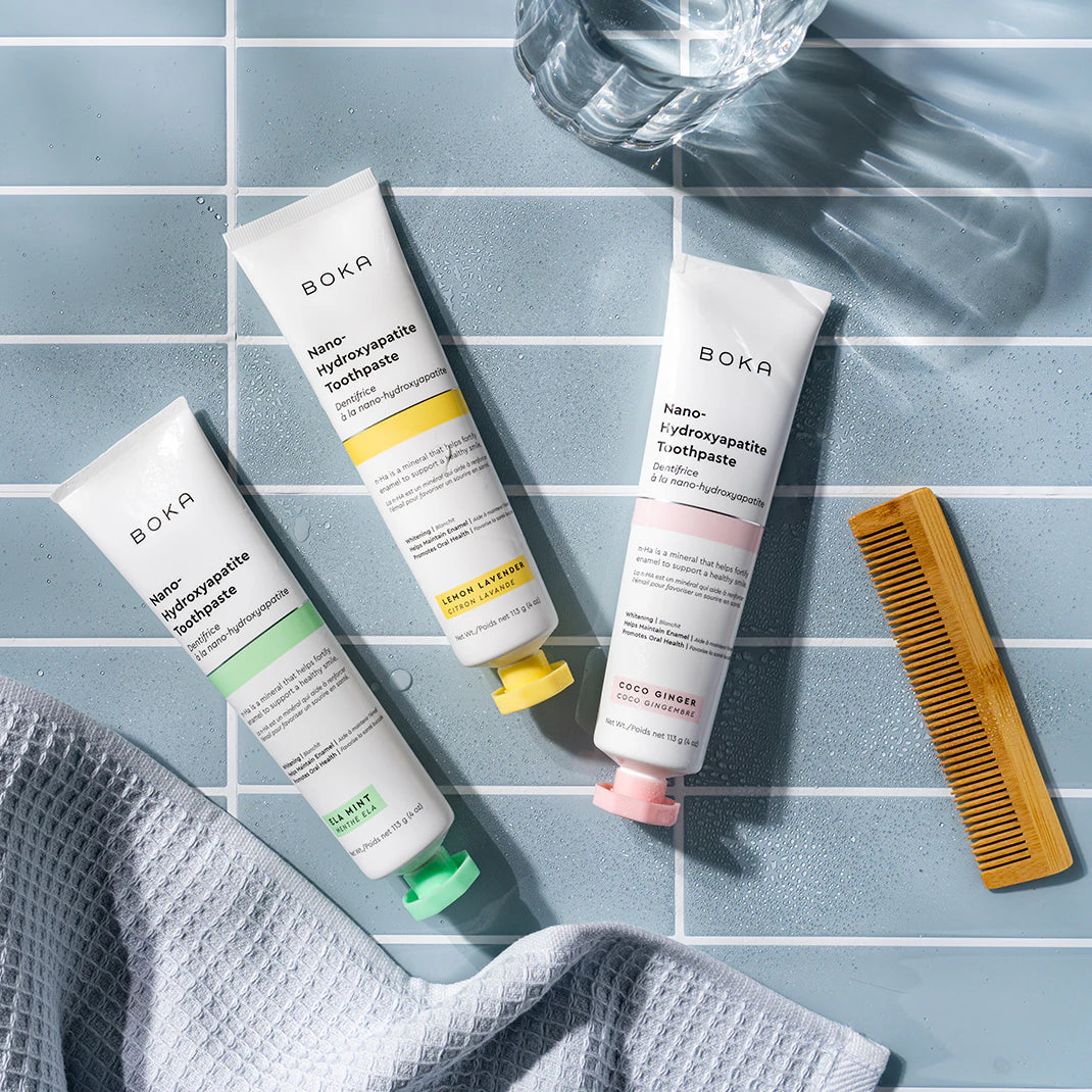 Three tubes of Boka toothpaste, a glass of water, a yellow comb, and a gray towel on blue tiles.