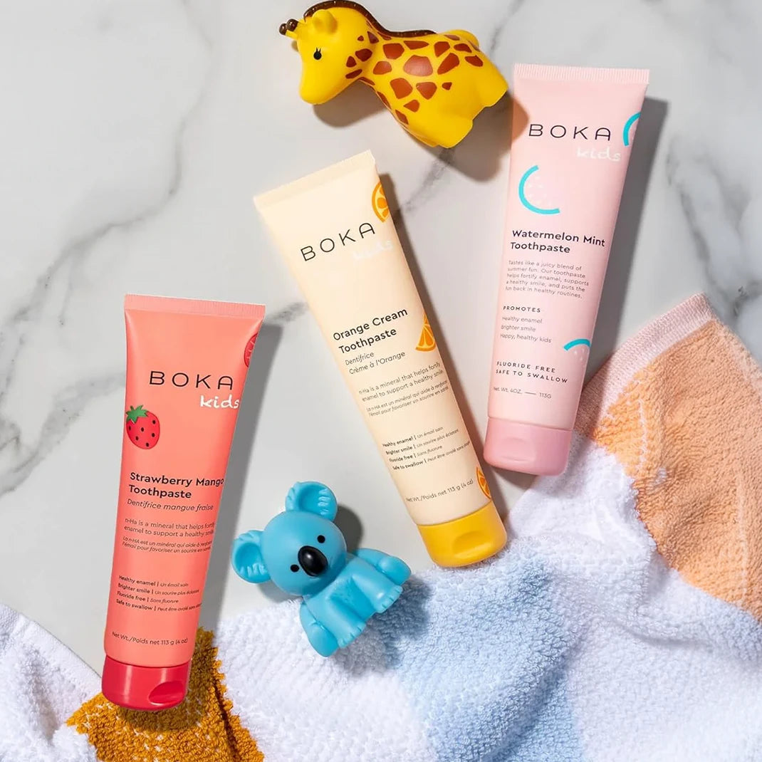 Three tubes of Boka kids toothpaste with a toy giraffe, toy koala, and a striped towel.