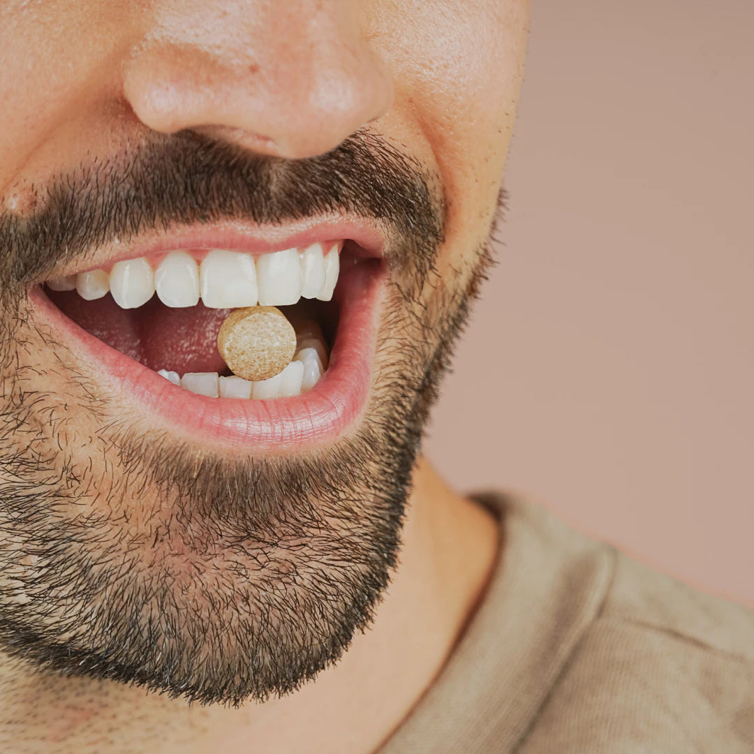 Close-up of a bearded man smiling with a round tablet resting between his teeth.