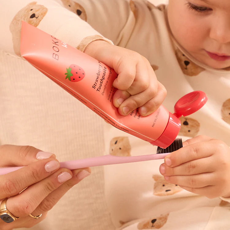 A child squeezes strawberry toothpaste onto a pink toothbrush held by an adult.