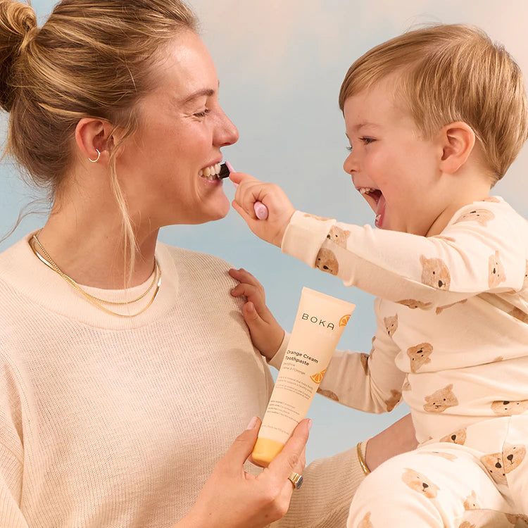 Smiling woman holds toothpaste as young child brushes her teeth with a pink toothbrush.