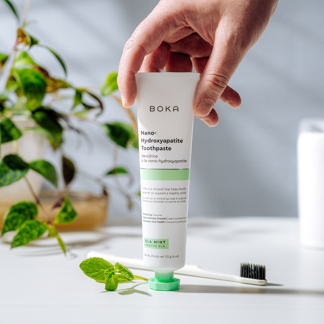 A hand holds a tube of Boka toothpaste above a toothbrush, with mint leaves and plants nearby.