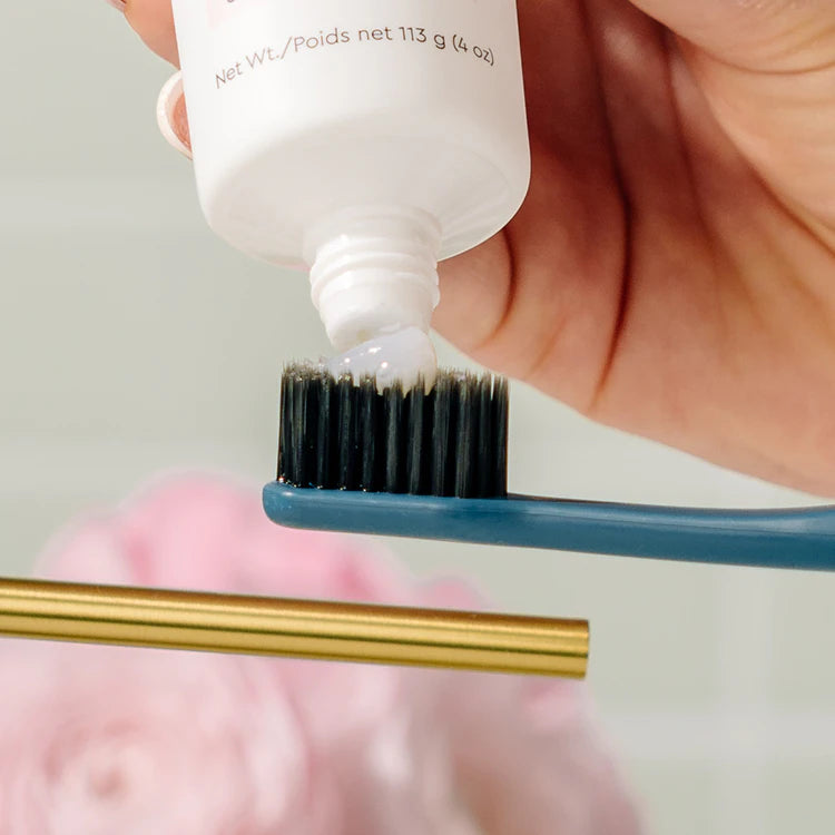 A hand squeezes toothpaste onto a blue toothbrush, with a blurred pink flower in the background.