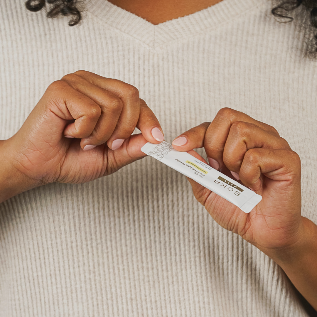 A person opens a small white packet with both hands, wearing a beige ribbed sweater.