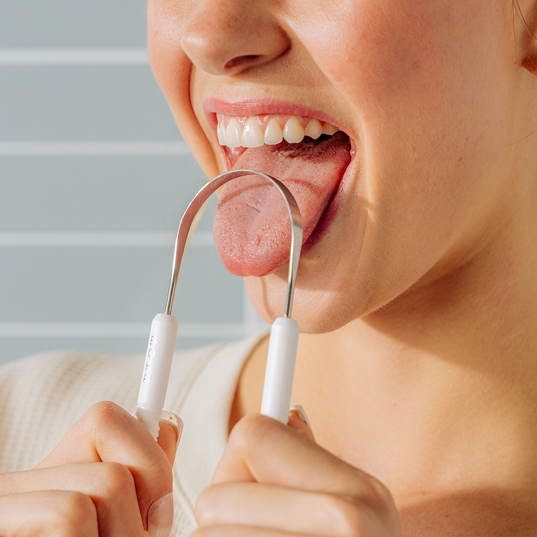 A woman uses a metal tongue scraper to clean her tongue, smiling with her mouth open.